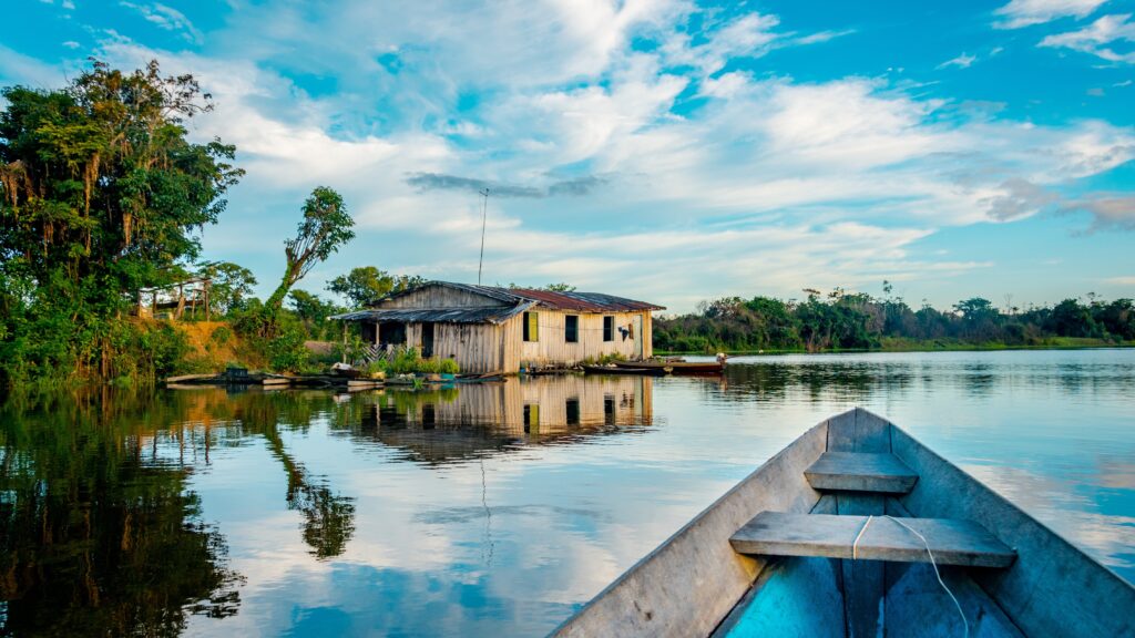 A canoe crosses a river in the Amazon rainforest beneath a blue sky.